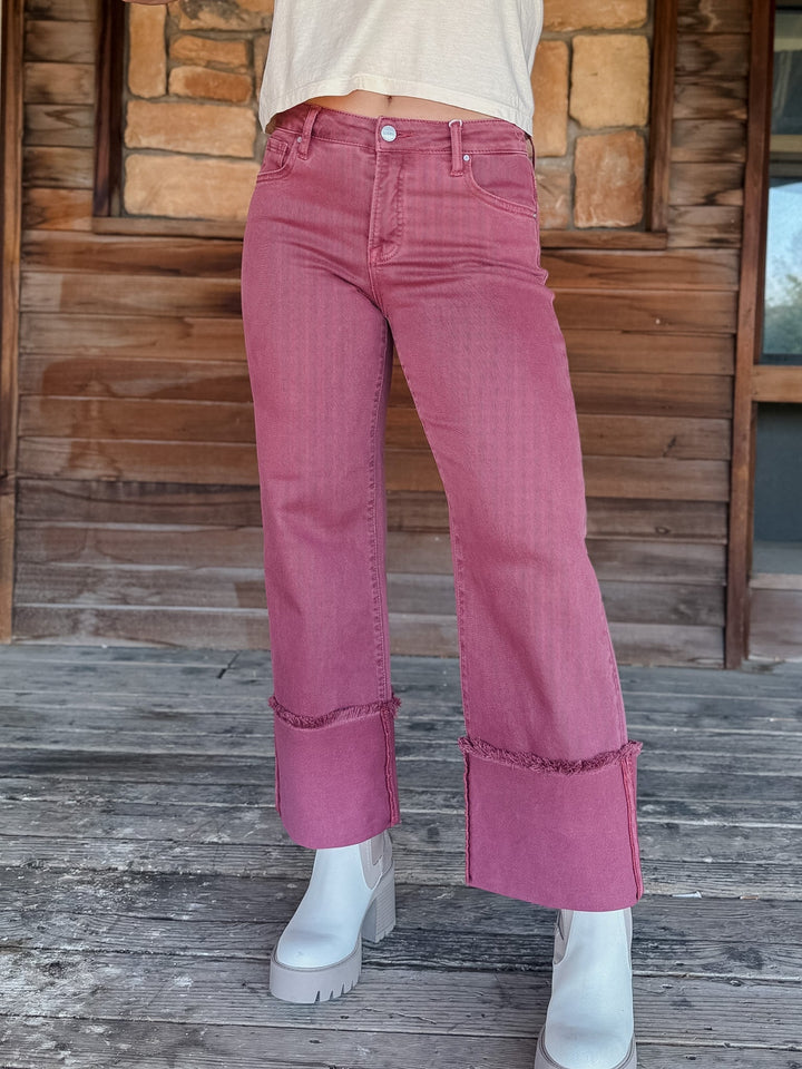 A person wearing dusty rose pink wide-leg cropped jeans with rolled cuffs, paired with white platform boots, standing on a weathered wooden deck in front of a rustic stone and wood wall.