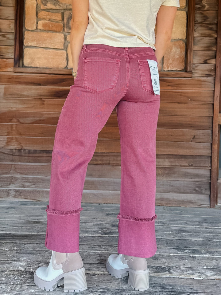 A person shown from the back wearing pink denim jeans with rolled cuffs, a cream t-shirt, and white platform shoes, standing in front of a rustic wooden and stone wall.