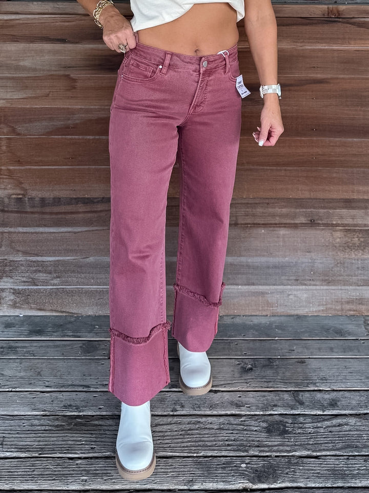 A person wearing dusty rose wide-leg cropped jeans with frayed hem details and rolled cuffs, paired with white ankle boots, standing against a rustic wooden plank wall.