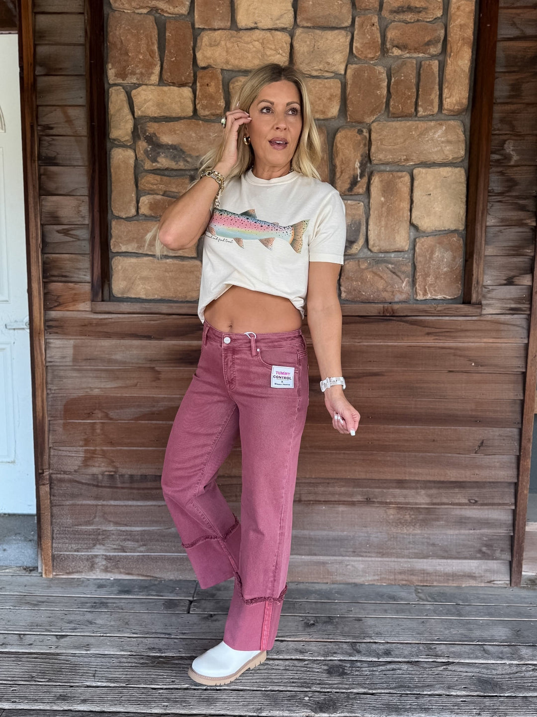 A woman stands on a rustic wooden porch in front of a stone wall, wearing a cream graphic t-shirt with a fish design and dusty rose wide-leg jeans.