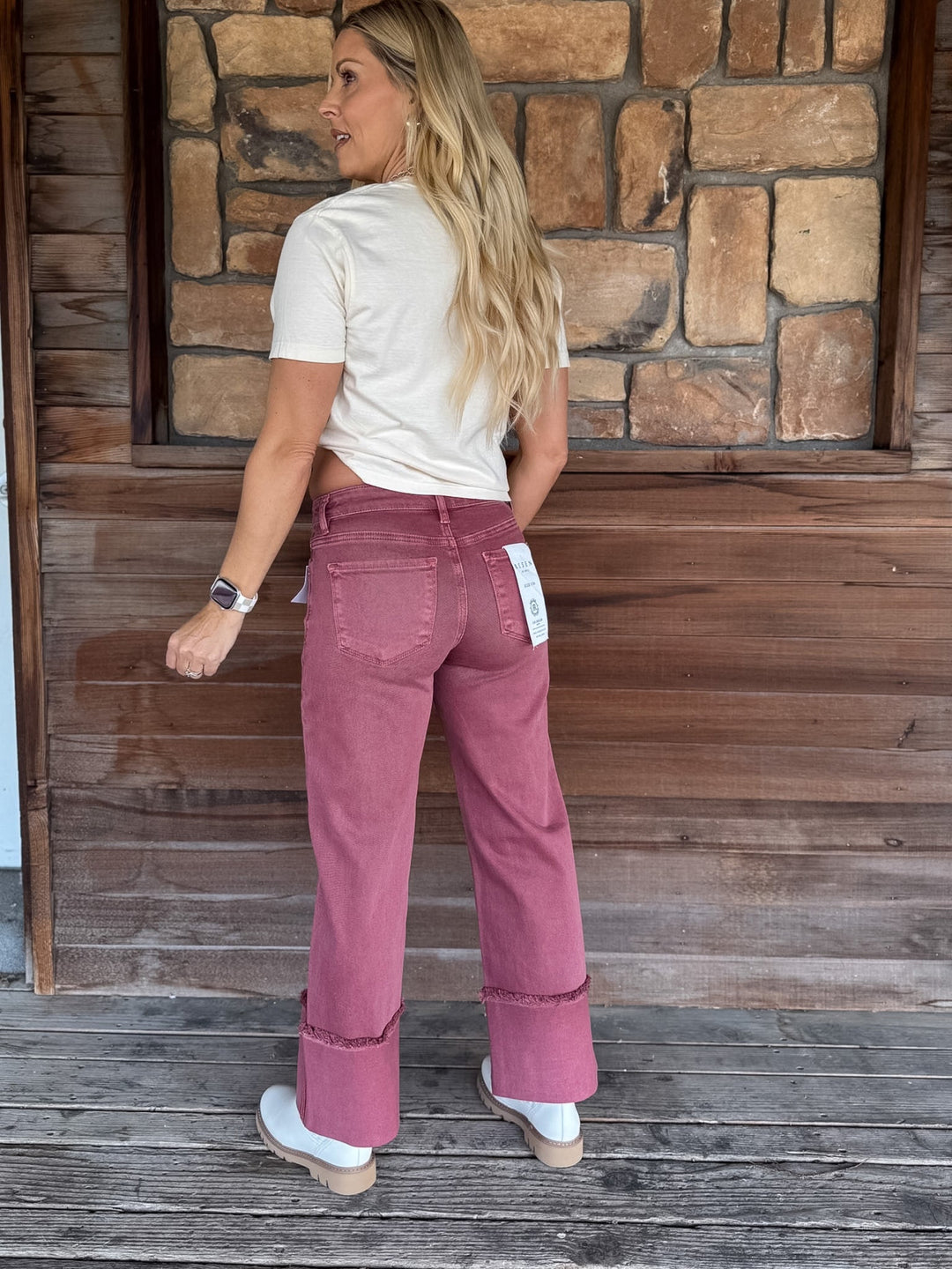 A woman stands on a wooden porch wearing a cream t-shirt and dusty rose wide-leg jeans with rolled cuffs, photographed from the back.