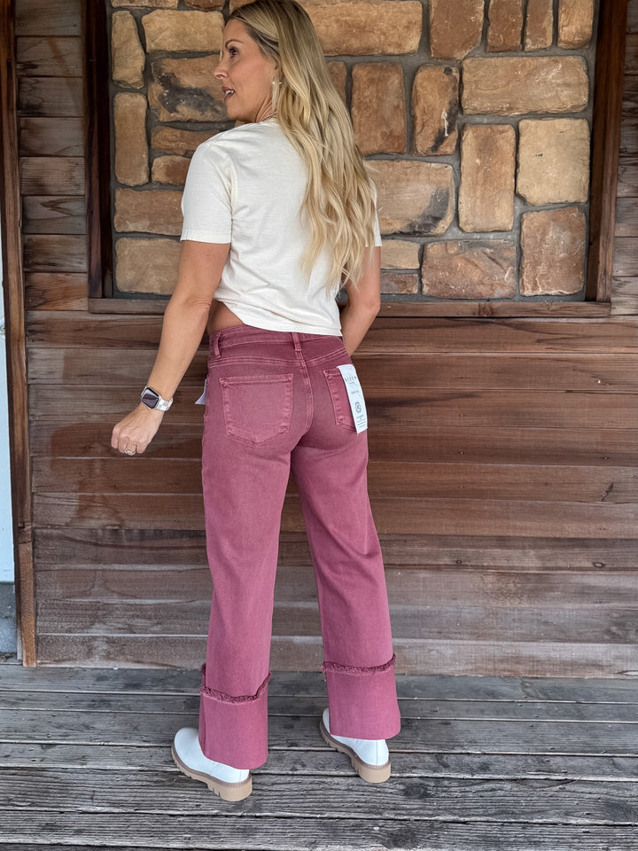 A woman stands on a wooden porch wearing a cream t-shirt and dusty rose wide-leg jeans with rolled cuffs, photographed from the back.