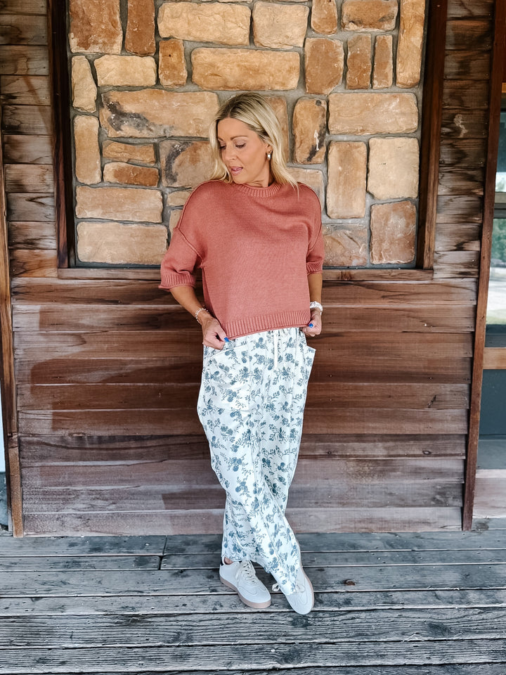 A woman stands on a wooden porch in front of a stone wall, wearing a dusty rose knit sweater, white pants with a blue floral print, and white sneakers.