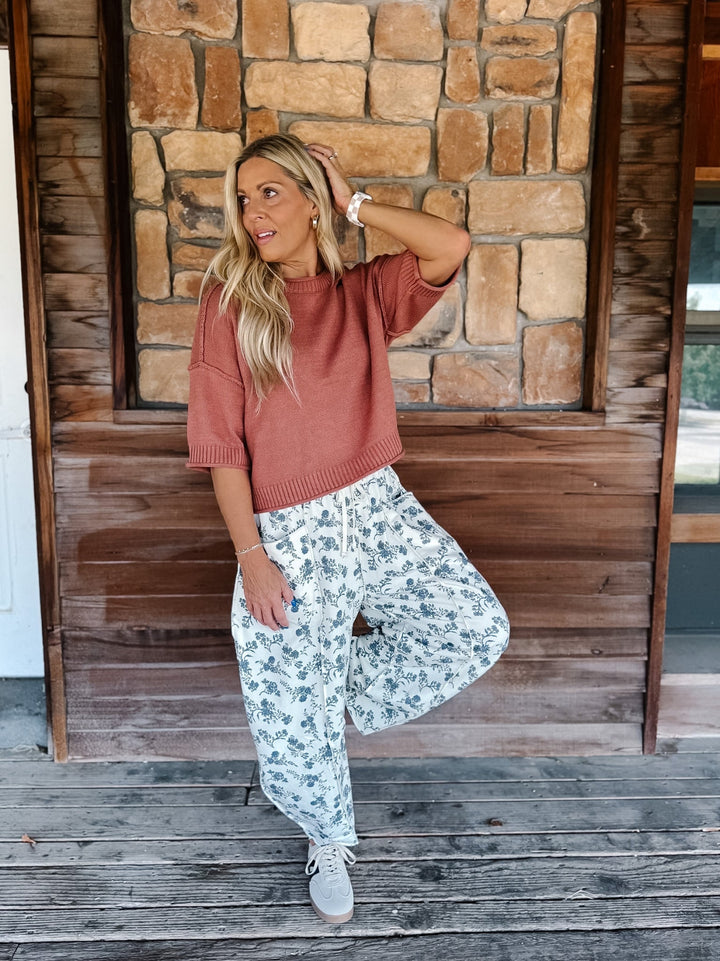 A woman posing on a rustic wooden porch in front of a stone wall, wearing a dusty rose short-sleeve knit top and white wide-leg sweatpants with a blue floral pattern.