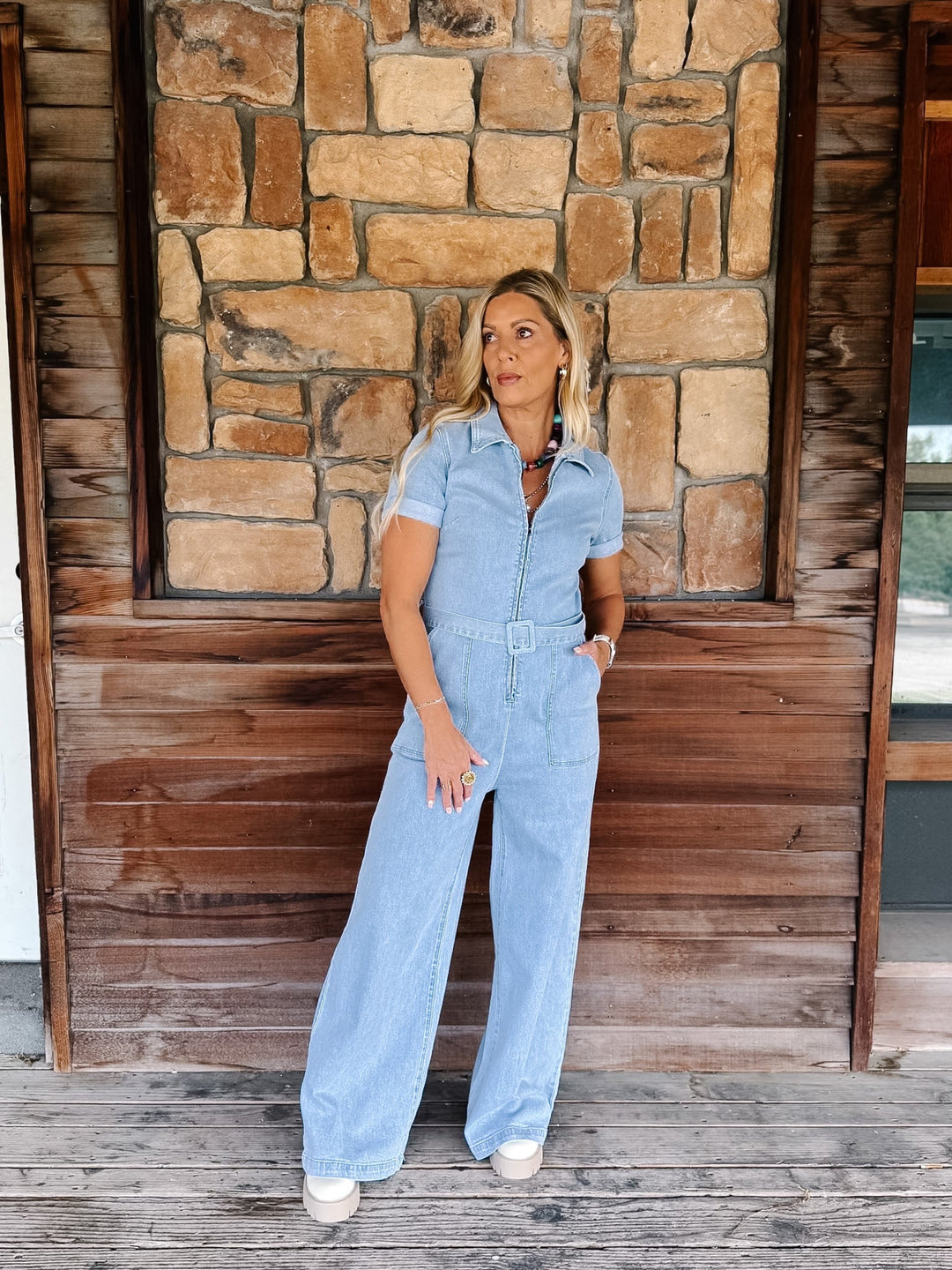 A woman modeling a light blue short-sleeve wide-leg jumpsuit with a matching belt, standing on a wooden porch in front of a stone wall.