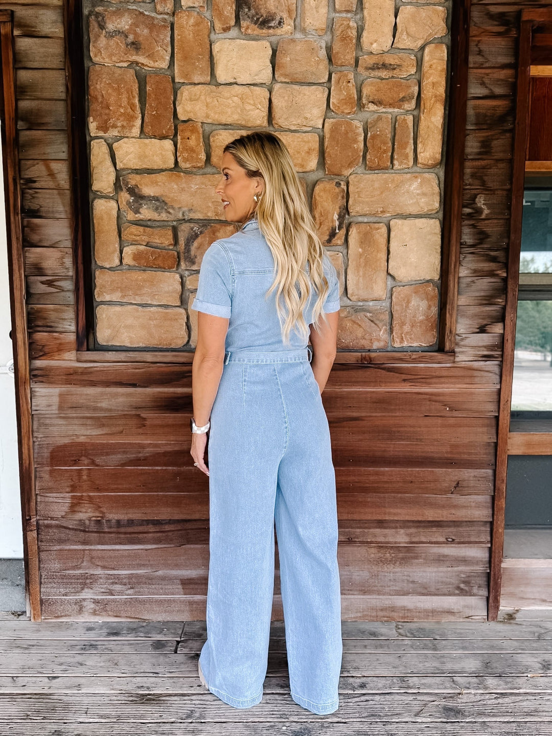 A woman stands with her back to the camera wearing a light blue denim short-sleeve jumpsuit with wide-leg pants, posed in front of a rustic stone and wood wall.