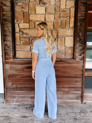 A woman stands with her back to the camera wearing a light blue denim short-sleeve jumpsuit with wide-leg pants, posed in front of a rustic stone and wood wall.