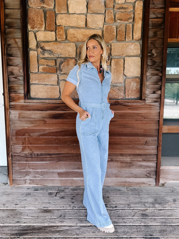 A woman modeling a light blue denim short-sleeve jumpsuit with a button-up front and wide legs, standing on a wooden porch in front of a stone and wood wall.