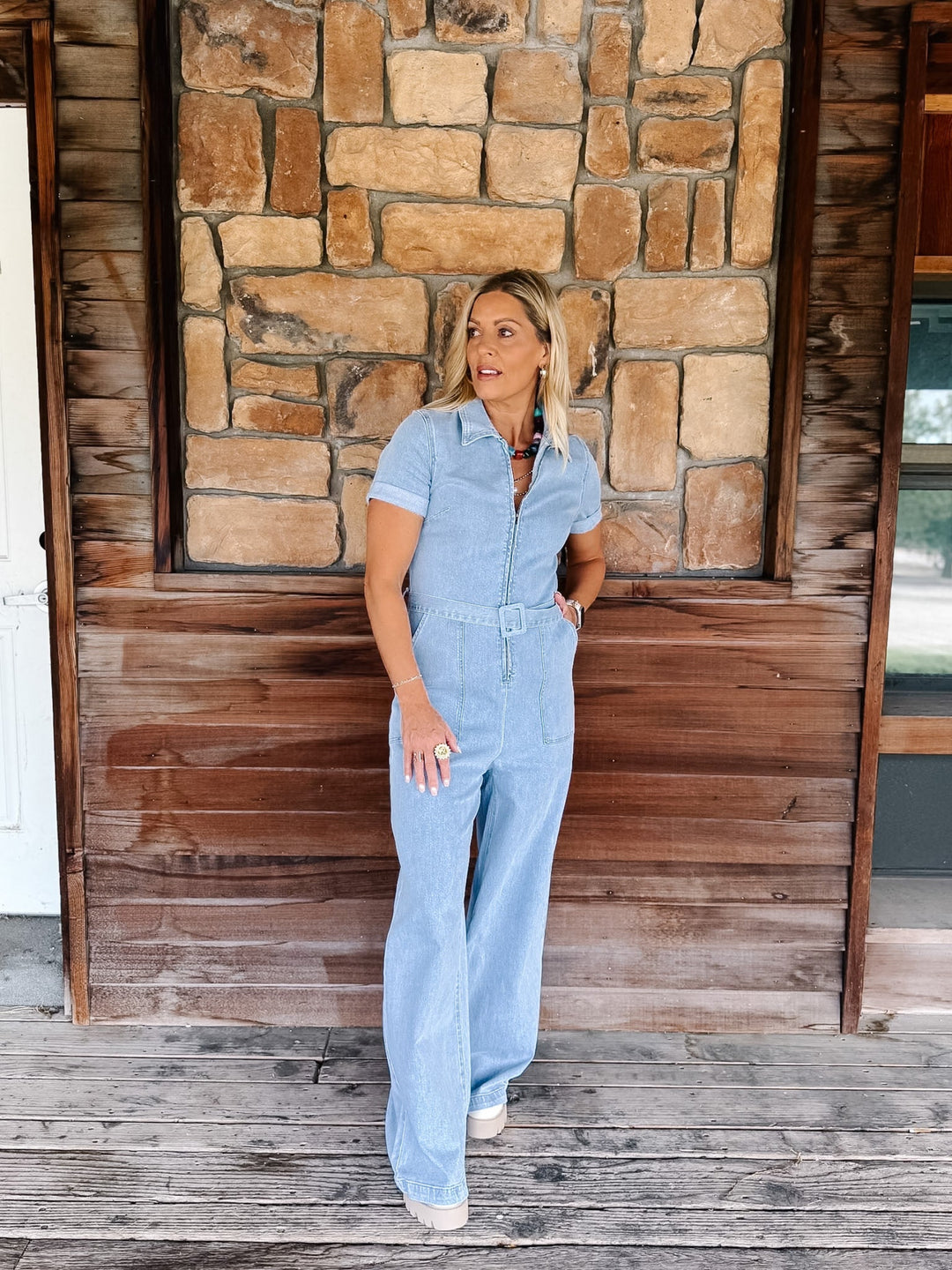 A woman wearing a light blue short-sleeve jumpsuit stands on a rustic wooden porch in front of a stone wall.