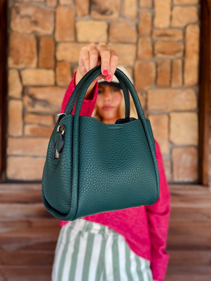 A woman holds up a dark teal pebbled leather handbag in front of her face, with a blurred stone wall and wooden floor in the background.