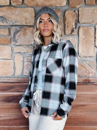 A woman wearing a black, gray, and white plaid flannel shirt jacket, a gray knit beanie, a cream pants, standing in front of a rustic stone wall.