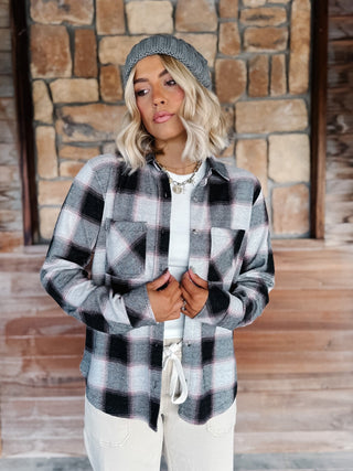 A woman wearing a black, gray, and white plaid flannel shirt jacket, a gray knit beanie, a cream pants, standing in front of a rustic stone wall.