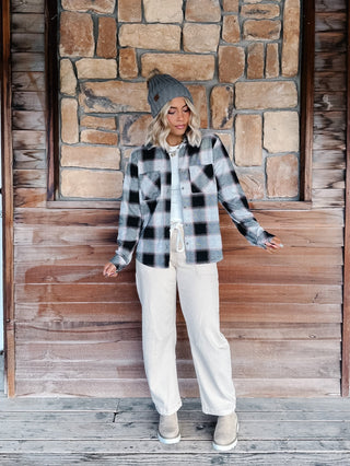A woman wearing a black, gray, and white plaid flannel shirt jacket, a gray knit beanie, a cream pants, standing in front of a rustic stone wall.