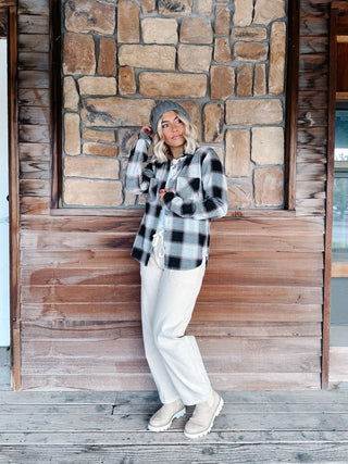 A woman wearing a black, gray, and white plaid flannel shirt jacket, a gray knit beanie, a cream pants, standing in front of a rustic stone wall.