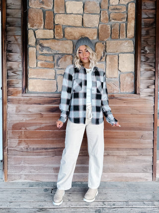 A woman wearing a black, gray, and white plaid flannel shirt jacket, a gray knit beanie, a cream pants, standing in front of a rustic stone wall.