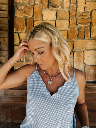 A woman wearing a light blue and white micro-dot patterned camisole top with a scalloped V-neckline,  standing in front of a rustic stone wall.