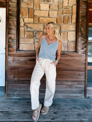 A woman wearing a light blue and white micro-dot patterned camisole top with a scalloped V-neckline, paired with off-white pants, standing in front of a rustic stone wall.