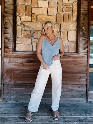 A woman wearing a light blue and white micro-dot patterned camisole top with a scalloped V-neckline, paired with off-white pants, standing in front of a rustic stone wall.