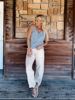 A woman wearing a light blue and white micro-dot patterned camisole top with a scalloped V-neckline, paired with off-white pants, standing in front of a rustic stone wall.
