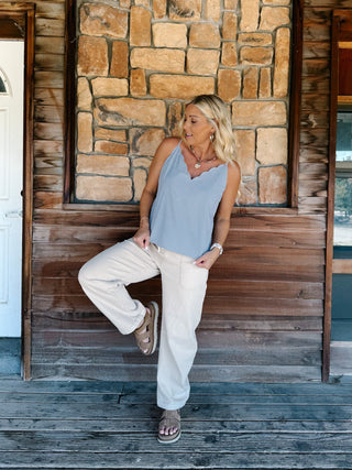 A woman wearing a light blue and white micro-dot patterned camisole top with a scalloped V-neckline, paired with off-white pants, standing in front of a rustic stone wall.