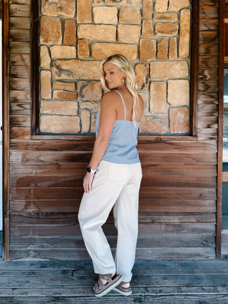 A woman wearing a light blue and white micro-dot patterned camisole top with a scalloped V-neckline, paired with off-white pants, standing in front of a rustic stone wall.