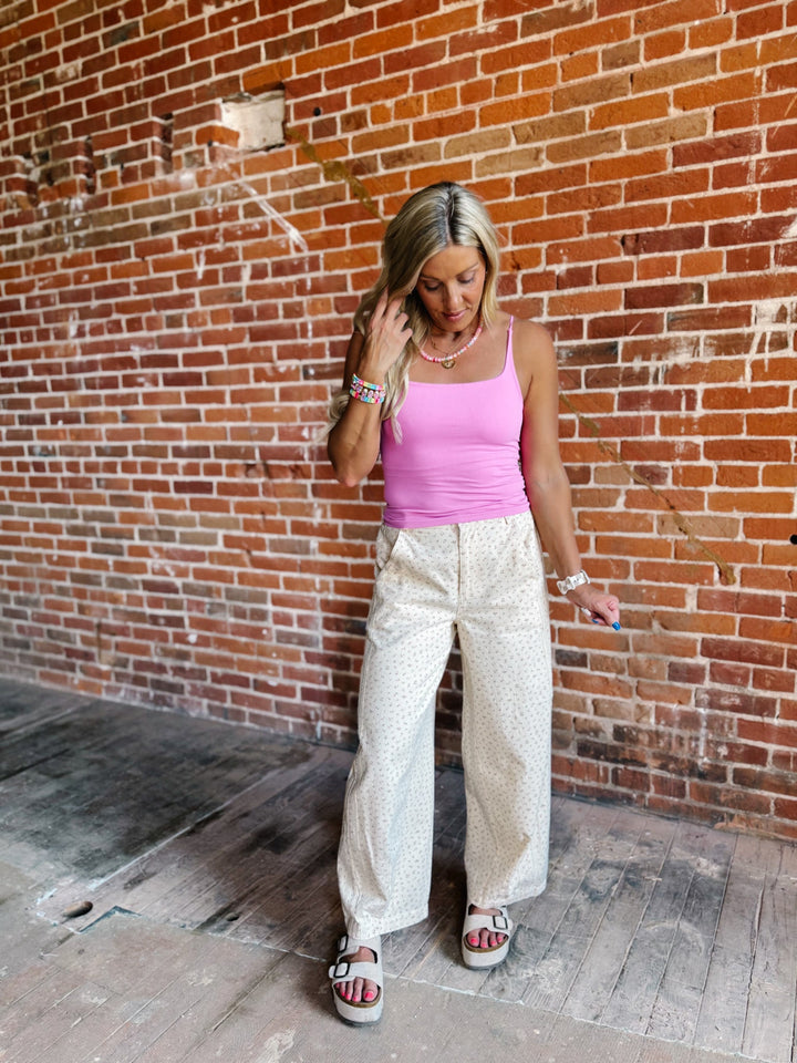 A woman stands against a red brick wall wearing a bright pink tank top, cream wide-leg pants with a subtle pattern, and white slide sandals.