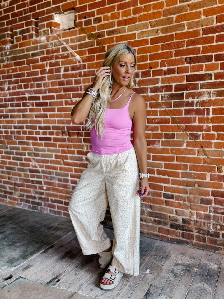 A woman posing in front of a brick wall wearing a pink tank top, white patterned wide-leg pants, and white platform sandals.