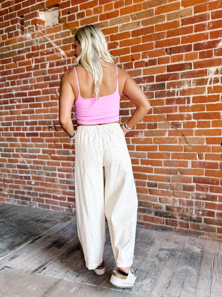 A woman stands with her back to the camera wearing a bright pink spaghetti-strap crop top and cream wide-leg pants with a tiny white pattern, set against a red-brown brick wall.