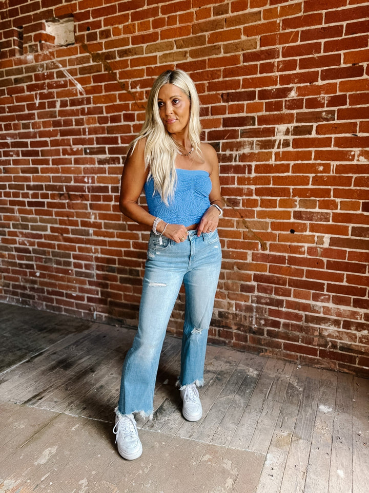 A woman with long blonde hair stands in front of a red brick wall wearing a bright blue strapless tube top, light-wash blue distressed jeans, and white sneakers.