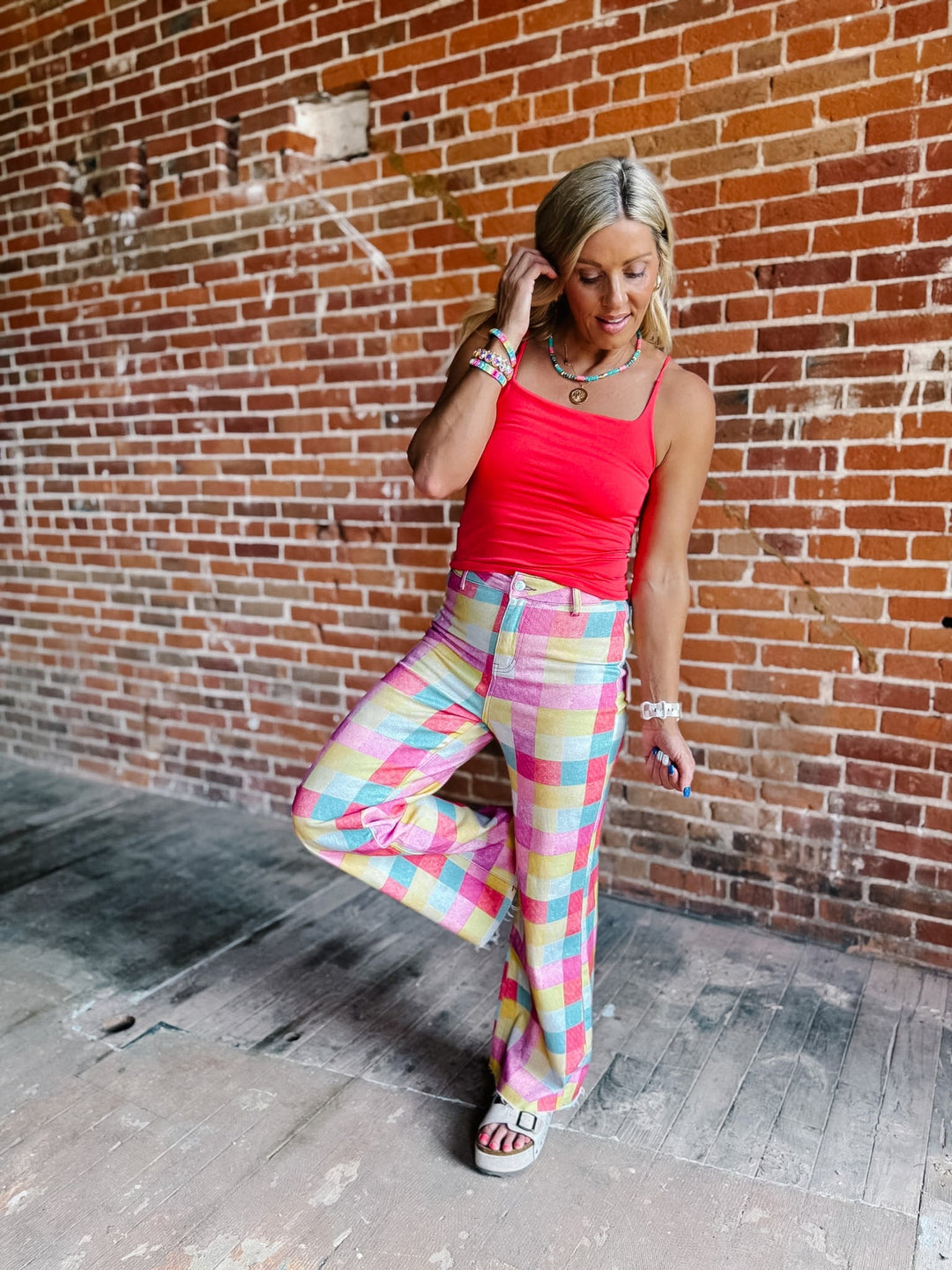 A woman posing in front of a brick wall wearing a bright coral red tank top and pastel multicolor plaid wide-leg pants, paired with white slide sandals.