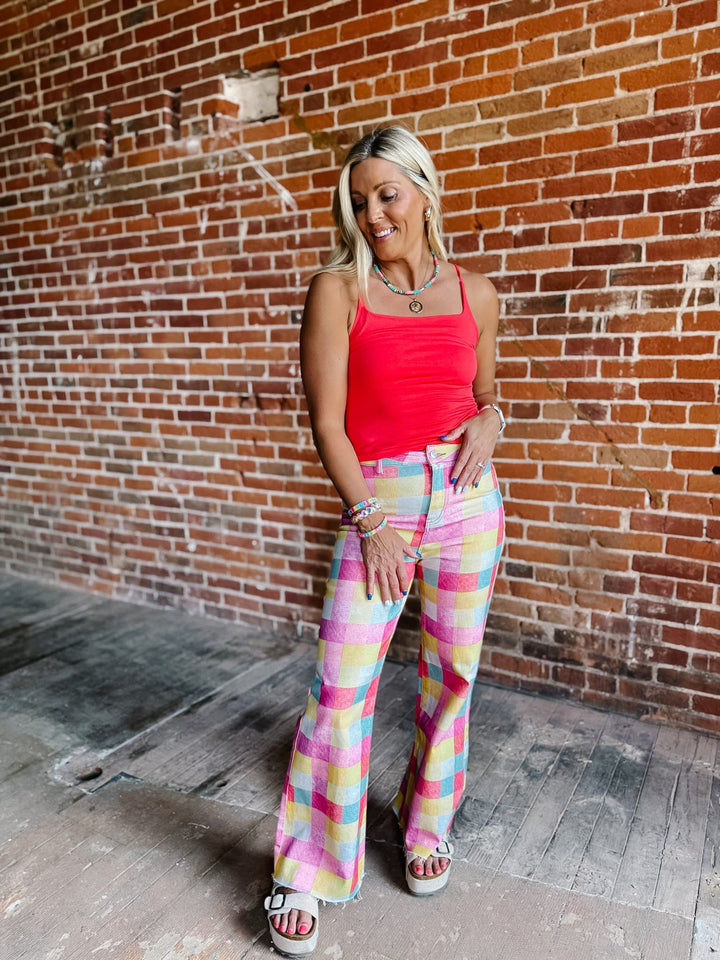 A woman posing in front of a brick wall wearing a bright coral pink ribbed tank top and pastel multicolor plaid wide-leg flare pants.