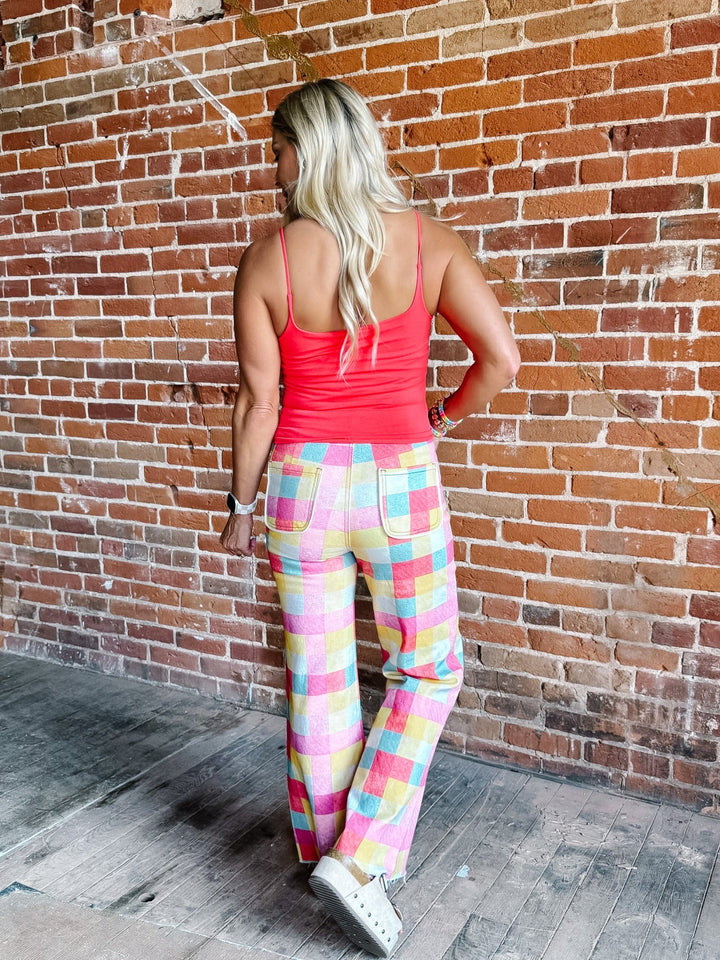 A woman stands with her back to the camera wearing colorful pastel plaid pants and a coral pink tank top, posing in front of a rustic red brick wall.
