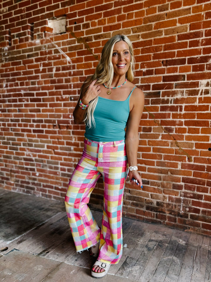 A woman posing in front of a red brick wall, wearing a teal tank top and colorful pastel plaid wide-leg pants.