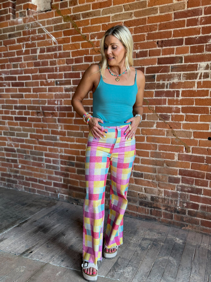 A woman posing in front of a brick wall wearing a bright turquoise tank top and colorful pastel plaid wide-leg pants.