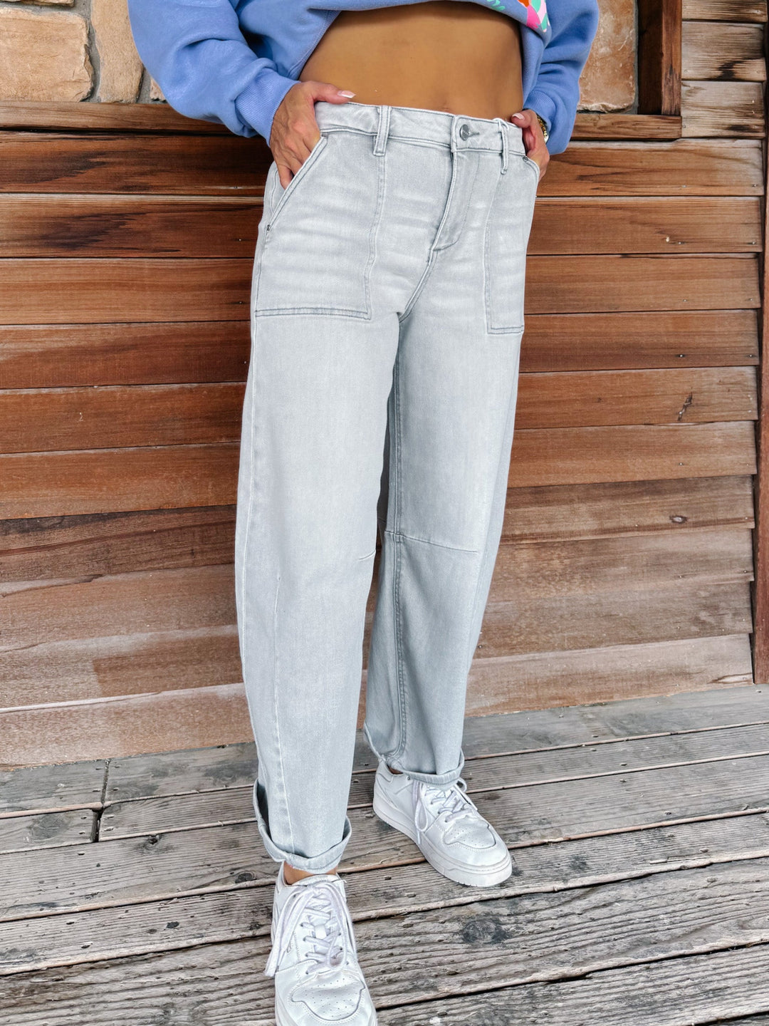 A person wearing light-wash, light-blue straight-leg denim jeans with front patch pockets, styled with a blue sweatshirt and white sneakers, standing against a rustic wooden wall.