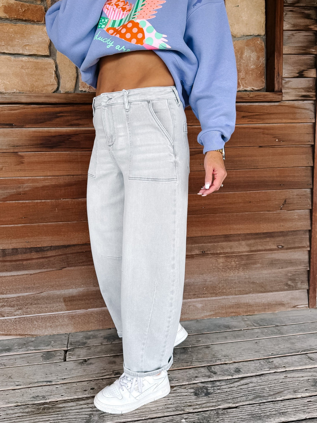 A person wearing light-wash, wide-leg denim jeans with a blue cropped sweatshirt and white sneakers, standing on a wooden deck in front of a wood-paneled wall.