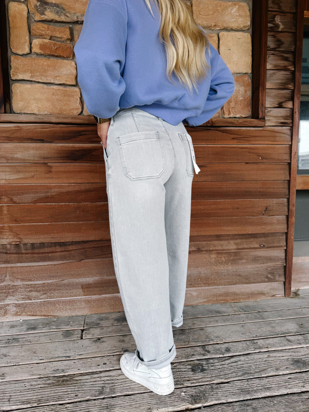A person standing on a wooden deck in front of a wood and stone wall, wearing light gray relaxed-fit jeans, a dusty blue sweatshirt, and white sneakers.