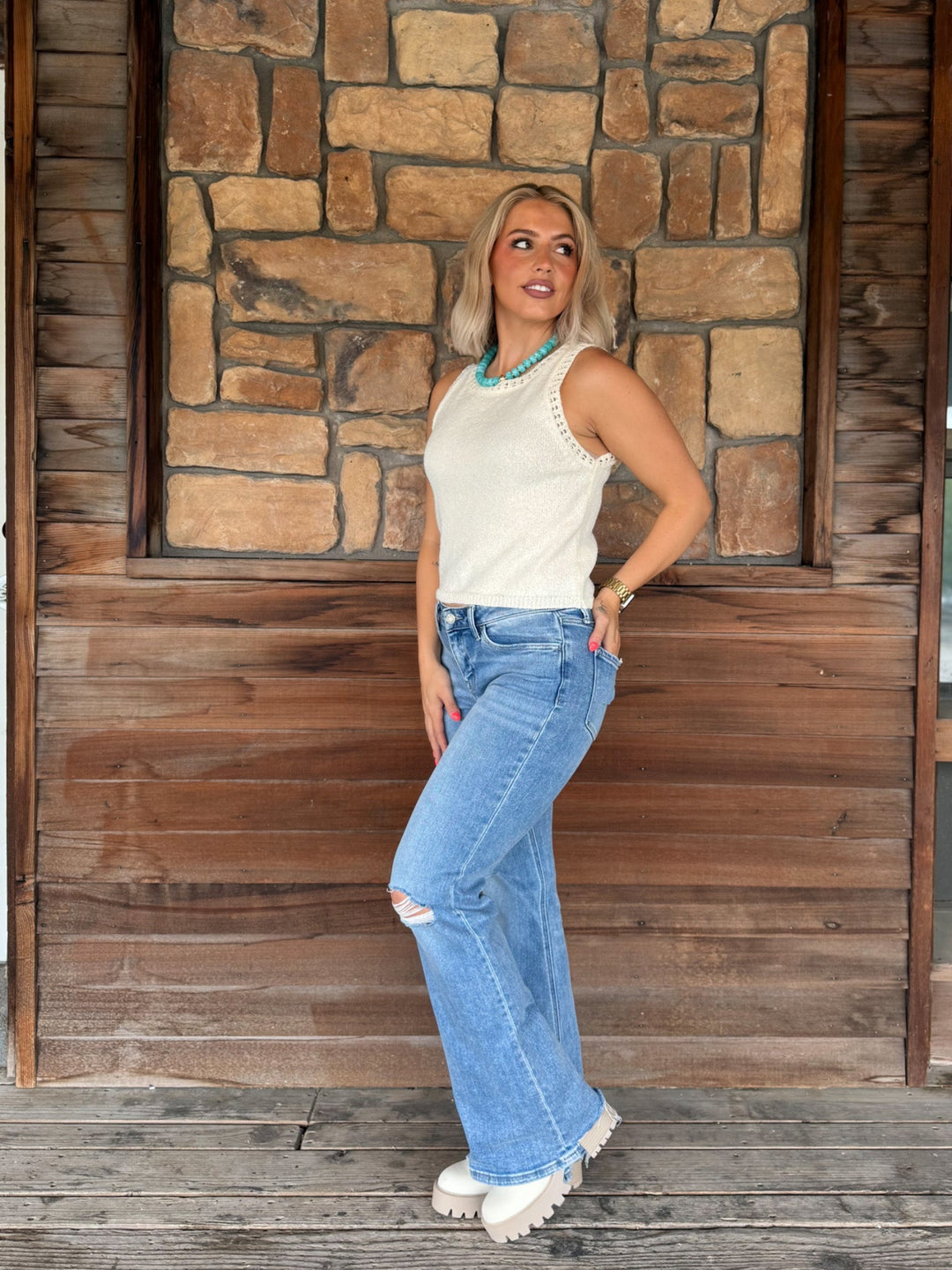 A woman modeling a cream sleeveless knit top and light blue distressed flare jeans with ripped knees, standing on a wooden porch in front of a stone wall.