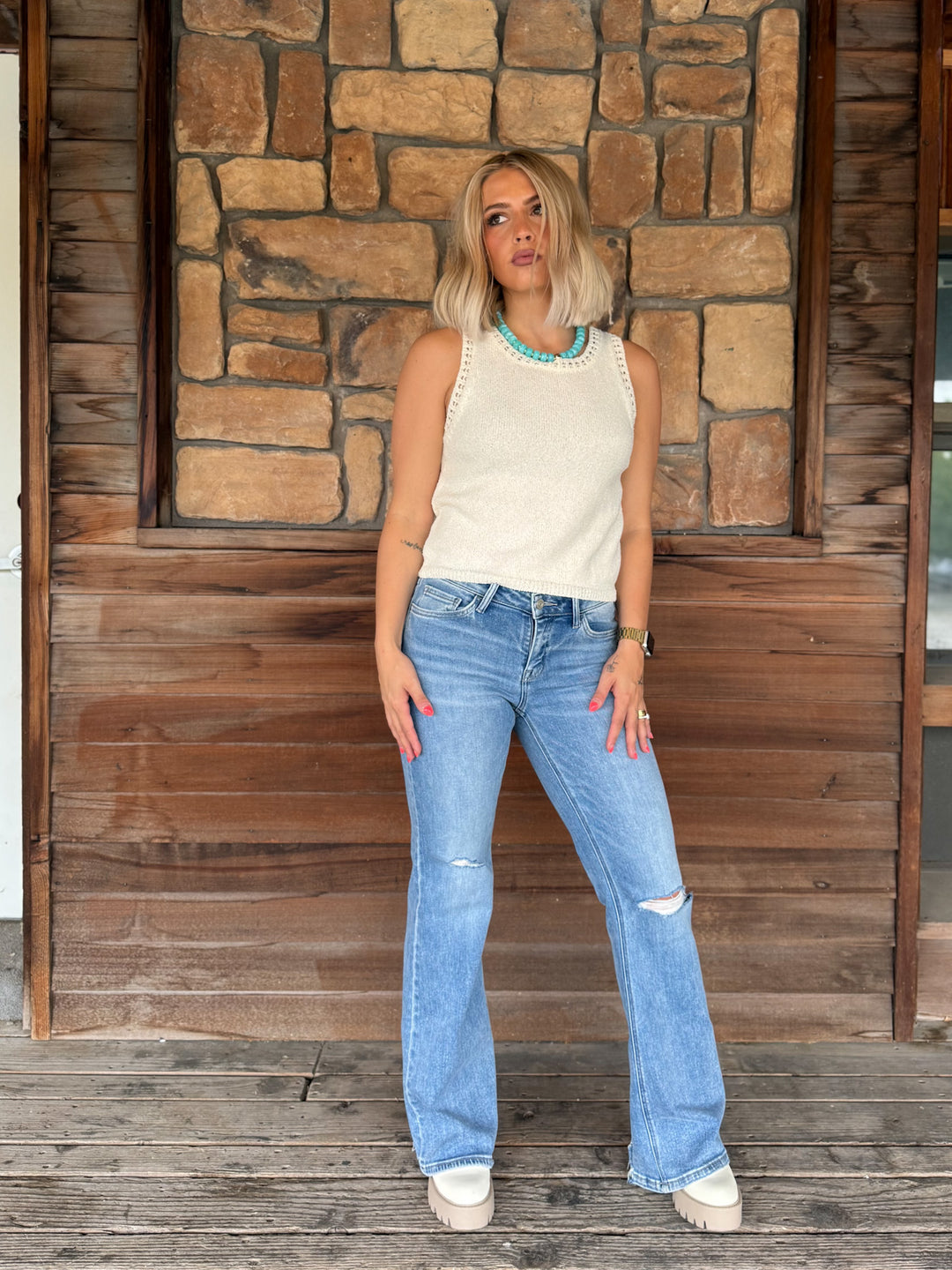 A woman stands on a wooden porch in front of a stone wall, wearing a cream sleeveless knit top with decorative trim, light-wash distressed blue jeans, and white platform sneakers.