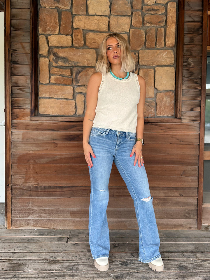 A woman stands on a wooden porch in front of a stone wall, wearing a cream sleeveless knit top with decorative trim, light-wash distressed blue jeans, and white platform sneakers.