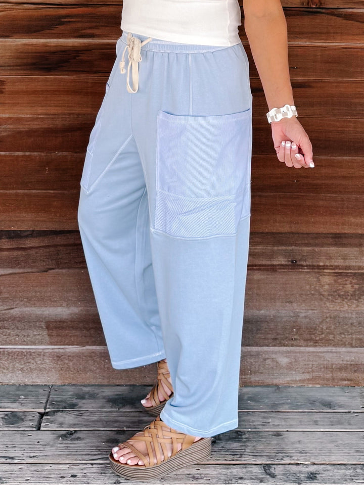 A person wearing light blue wide-leg drawstring pants with large front patch pockets, paired with a white top and tan wedge sandals, standing against a rustic wooden wall and deck.