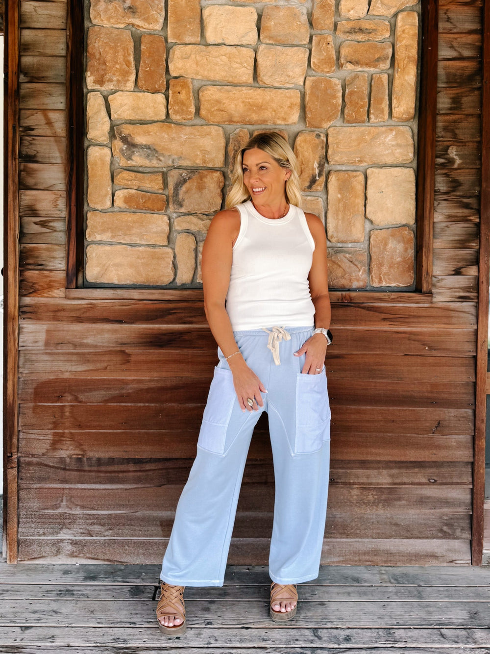 A woman stands on a wooden porch in front of a stone wall, wearing a white sleeveless top and light blue wide-leg pants with front patch pockets.