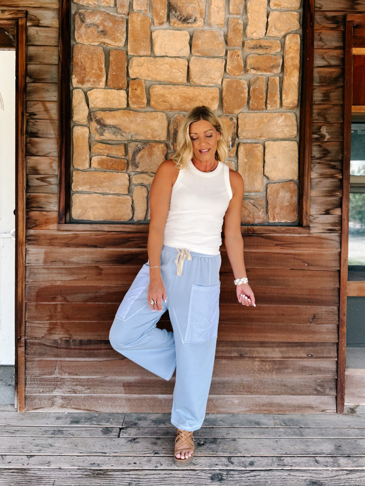 A woman posing on a rustic wooden porch in front of a stone wall, wearing light blue wide-leg drawstring pants and a white sleeveless top.