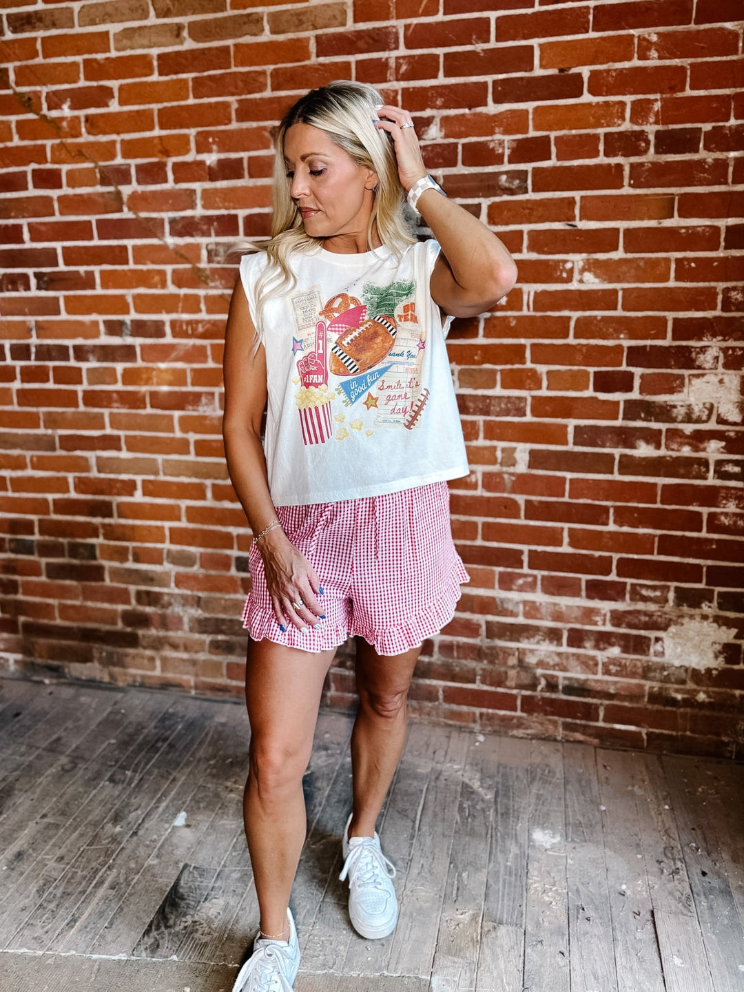 A woman stands in front of a brick wall wearing a white sleeveless graphic top, pink and white gingham shorts, and white sneakers.