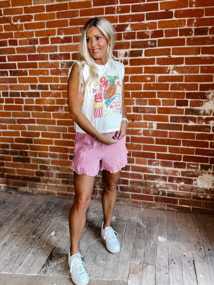 A woman stands in front of a red brick wall wearing a white sleeveless crop top with a colorful ice cream graphic, pink gingham ruffle shorts, and white sneakers.