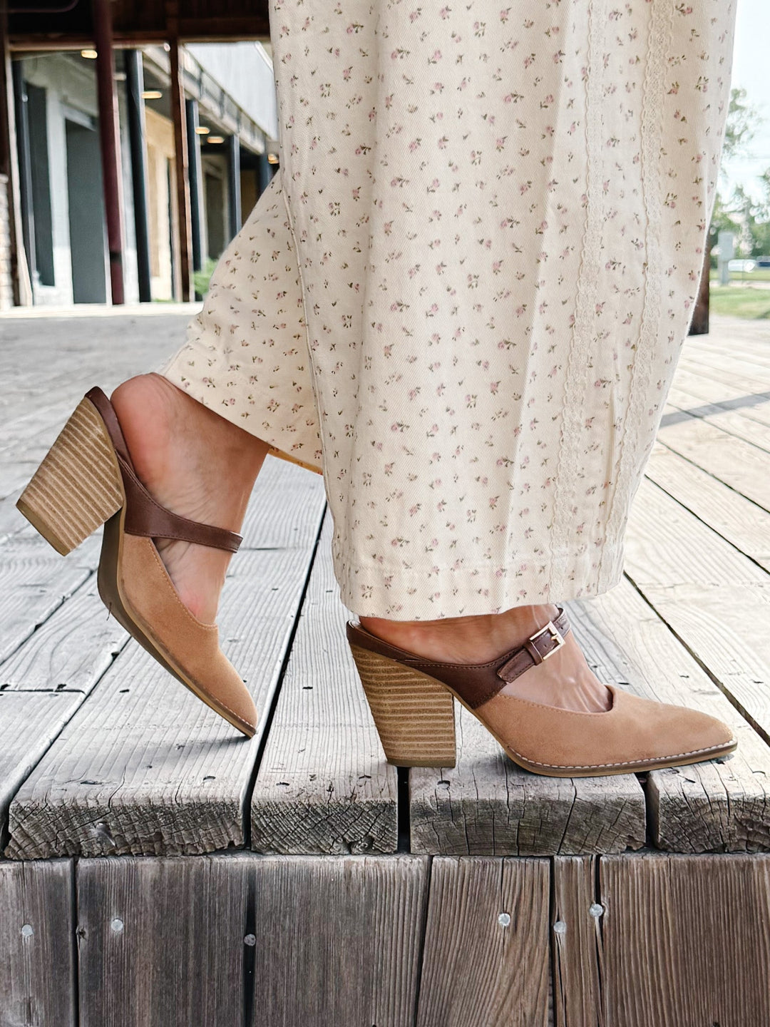A person wearing beige block-heel mule sandals with brown straps and a cream floral skirt, standing on a weathered wooden deck.