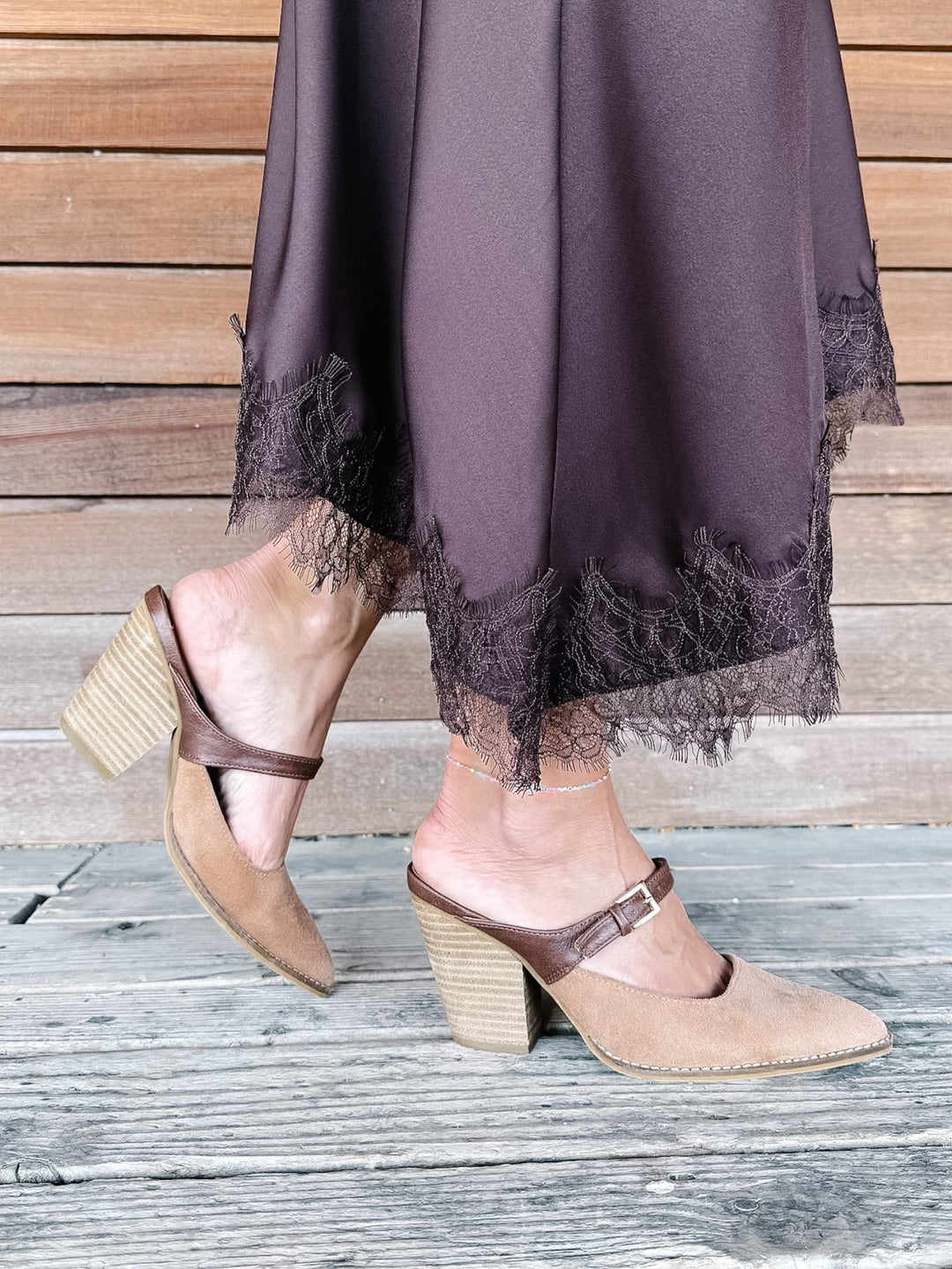 A person wearing taupe suede mule heels with brown straps and stacked block heels, paired with a dark brown skirt featuring lace trim, standing on a weathered wooden deck.