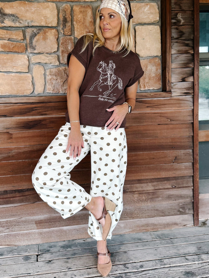 A woman posing on a rustic wooden porch wearing a brown graphic t-shirt, white pants with brown polka dots, and a beige headband.