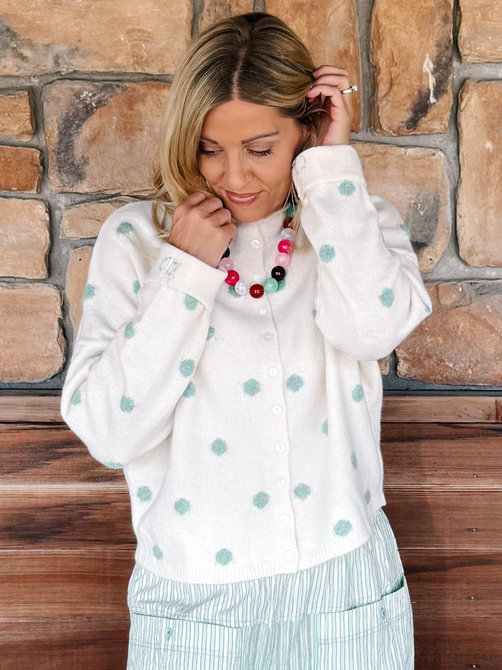 A woman wearing a white button-up cardigan with mint green polka dots, paired with a colorful beaded necklace and light green striped pants, standing in front of a rustic stone wall.