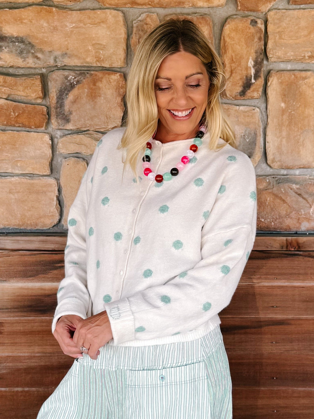 A woman wearing a cream zip-up cardigan with mint green polka dots and a striped skirt, standing in front of a rustic stone wall.